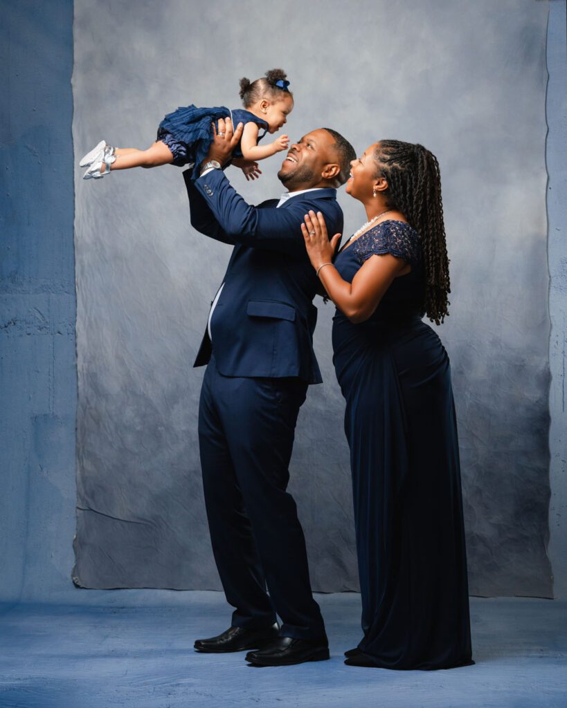 Father wearing a blue suit holds his baby girl wearing a blue dress in the air as mom in a blue gown looks at her daughter. 