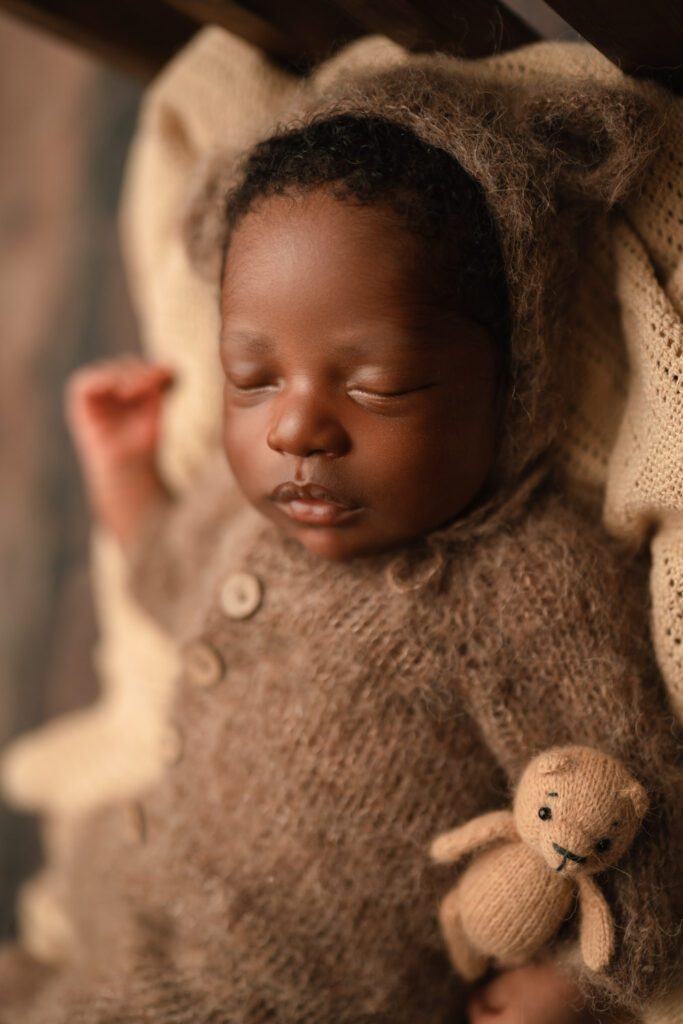 Sleeping newborn baby wearing a brown knit onesie and matching bear bonnet with ears holds a tan crochet bear stuffy.