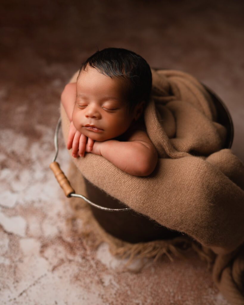 Baby boy with black hair rests his head on his hands while in a metal bucket filled with a soft brown blanket.