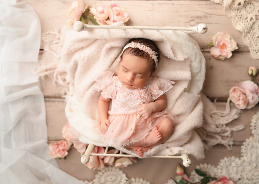Baby girl sleeps on her back on a white iron rod bed wearing a pink dress surrounded by flowers above a white wood floor. 