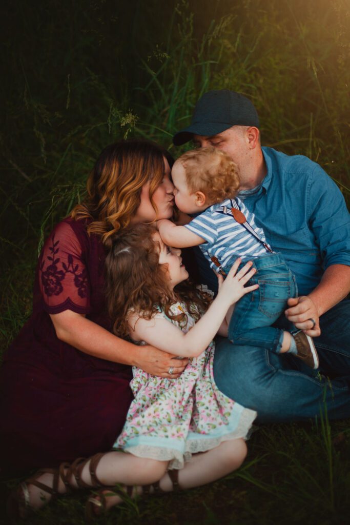 family of four laying down in grassy meadow snuggling closely and kissing baby boy