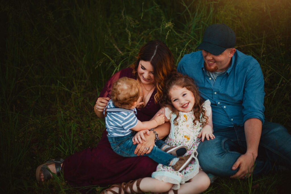 mom, dad and their two children sitting down close together in a meadow laughing 