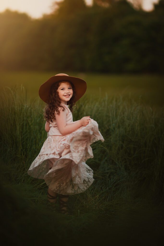 young girl with red hair in brimmed hat twirling her long ruffled dress in a meadow at sunset