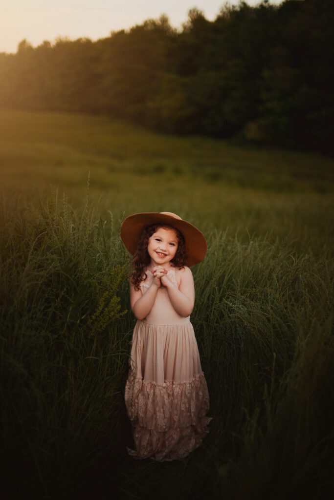 Young daughter in a blush dress and wide-brim hat standing alone in tall meadow grass.