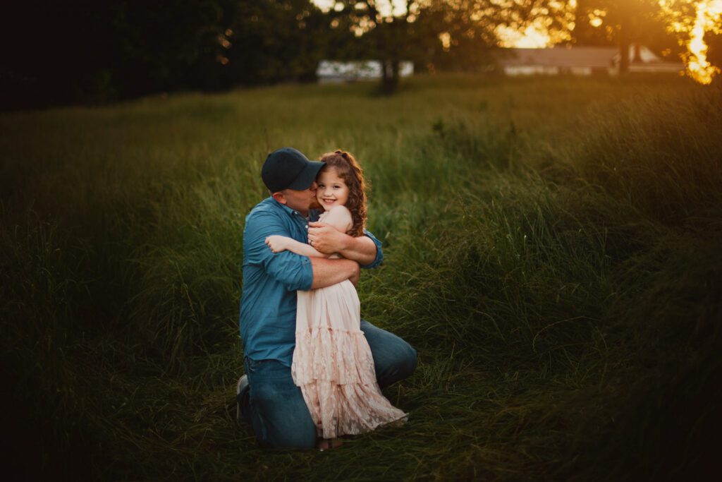 Dad kneeling in tall grass while hugging his smiling daughter close beside him.