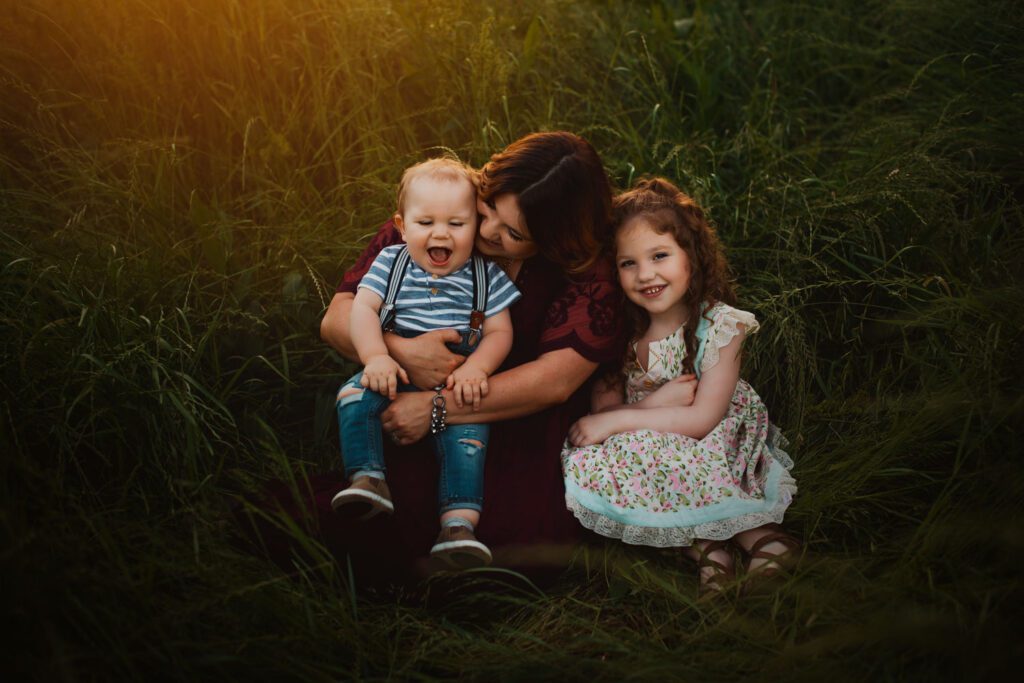 Mom sitting in tall grass with her baby son and young daughter gathered closely beside her.