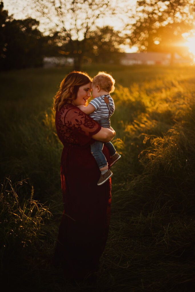 Mom holding her baby son close in tall grass with bright golden sunset light behind them.