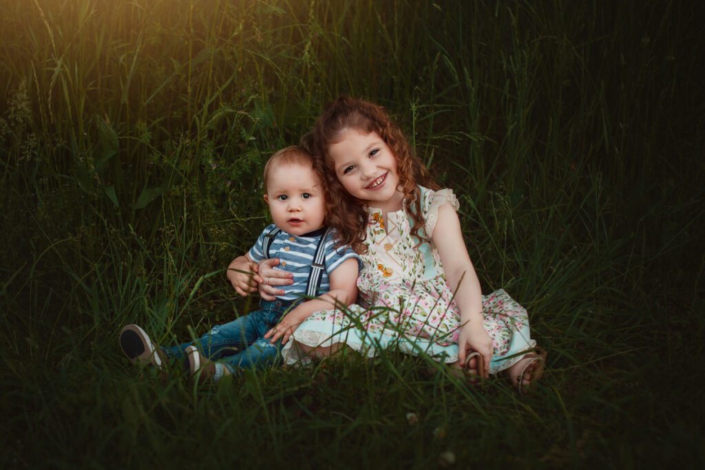 Brother and sister sitting close together in the grass with warm evening light behind them.
