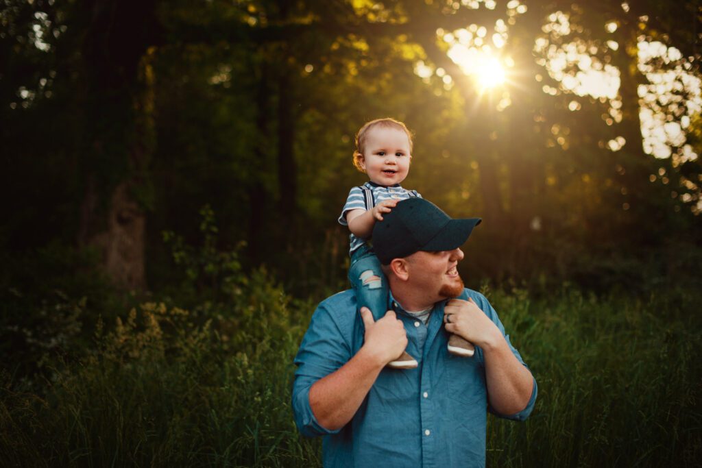 Dad carrying his baby son on his shoulders in tall grass with golden sunlight filtering through the trees.