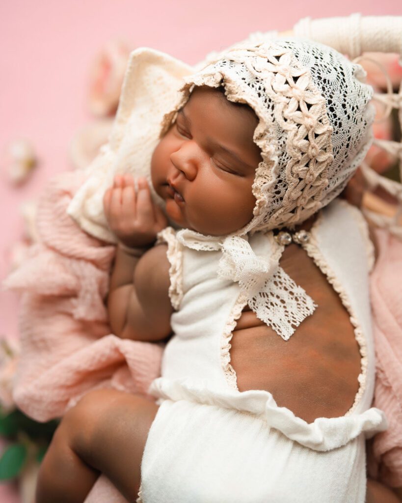Profile of a newborn baby girl in a cream lace bonnet, showing her cheeks against a soft pink backdrop.