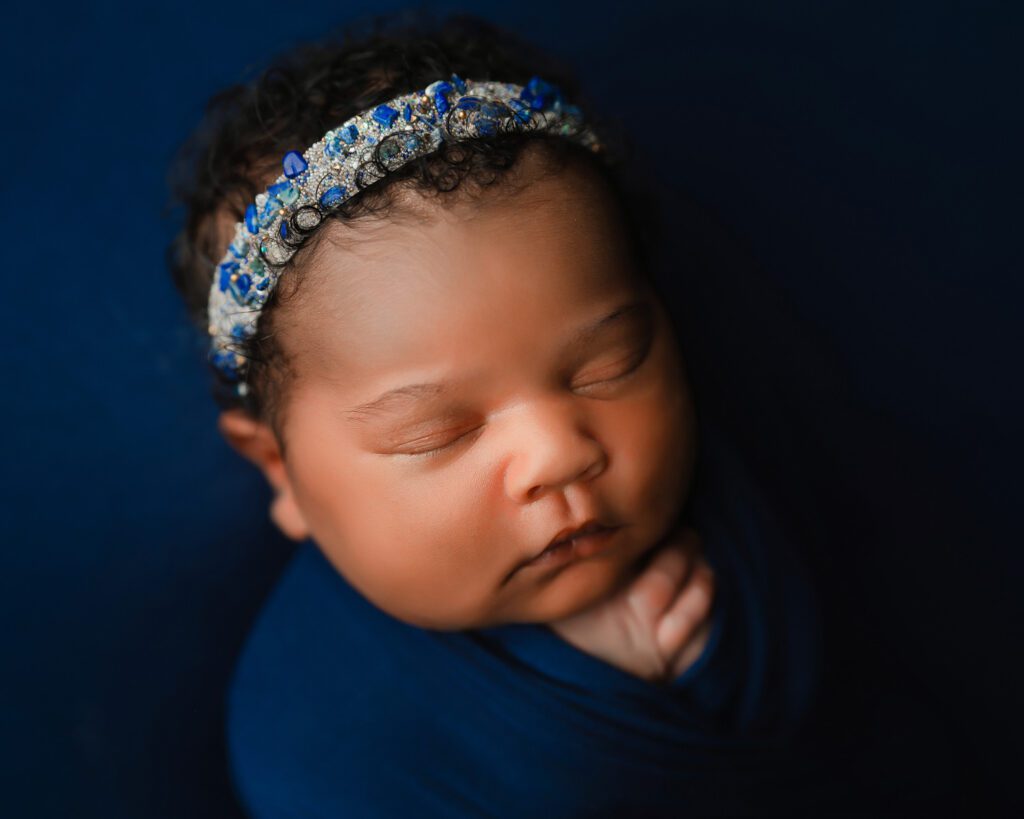 Close-up of a sleeping newborn baby girl in a navy swaddle with a blue crystal headband.