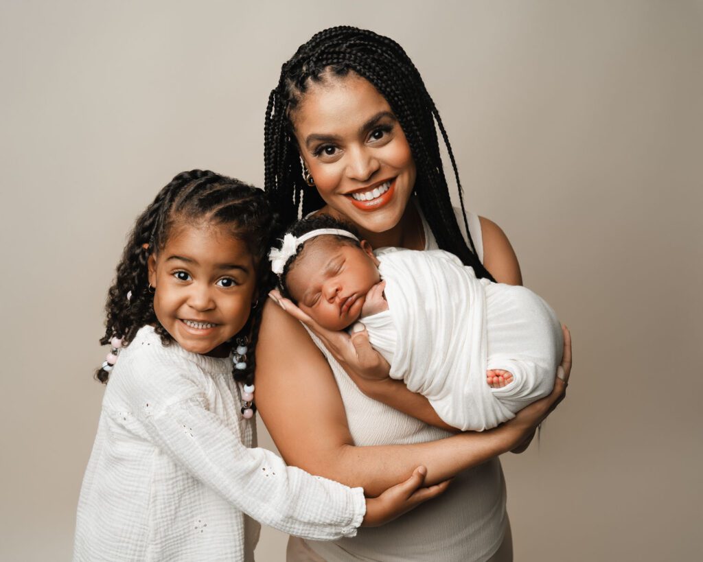 Mom holding her newborn daughter while big sister hugs close against a neutral studio backdrop.