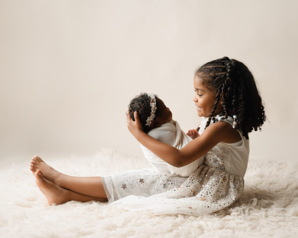 Big sister in a white sparkly dress holding her newborn baby sister during a Marietta newborn photographer session.