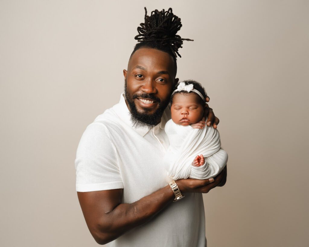 Dad smiling while holding his swaddled newborn daughter against a simple neutral backdrop.