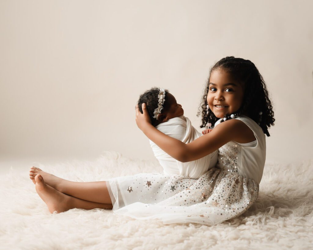 Big sister sitting on a furry rug holding her newborn sister, both dressed in white for a Marietta newborn photographer.
