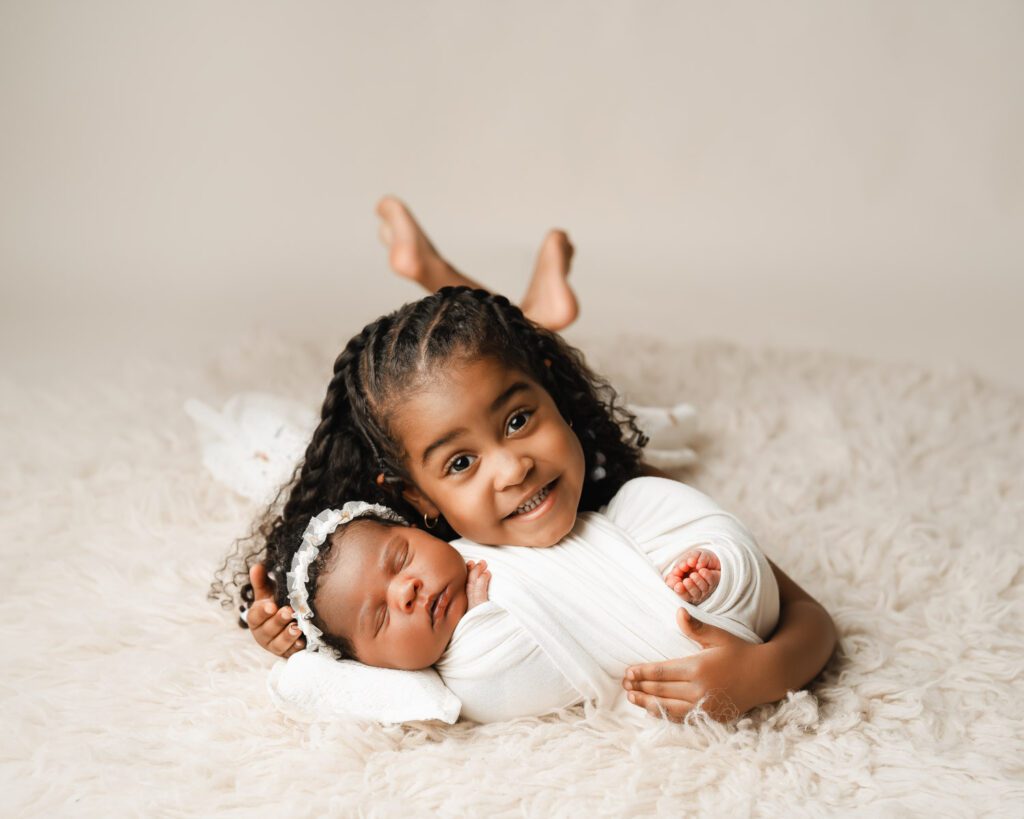 Big sister lying on a cream fur rug while holding her newborn sister wrapped in a soft white swaddle.