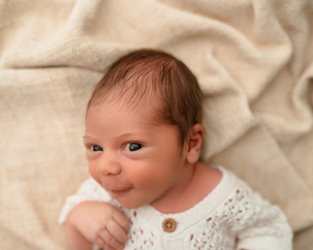 9 day old awake newborn boy wearing a white knit outfit on a tan blanket. 