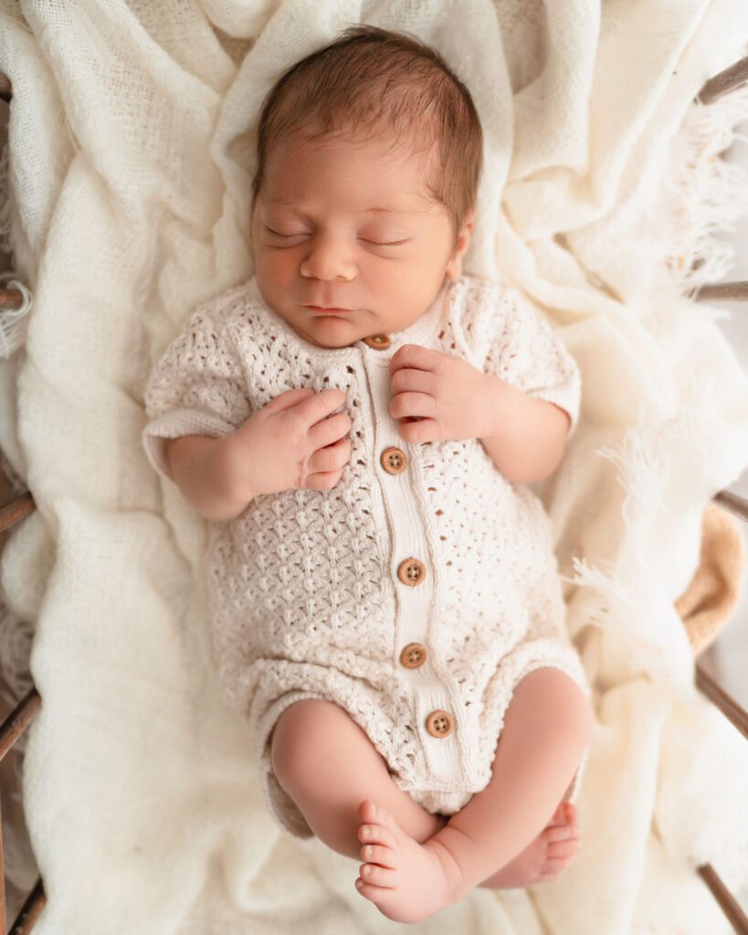 newborn baby wearing a white onesie while laying on white blankets in an antique crib.
