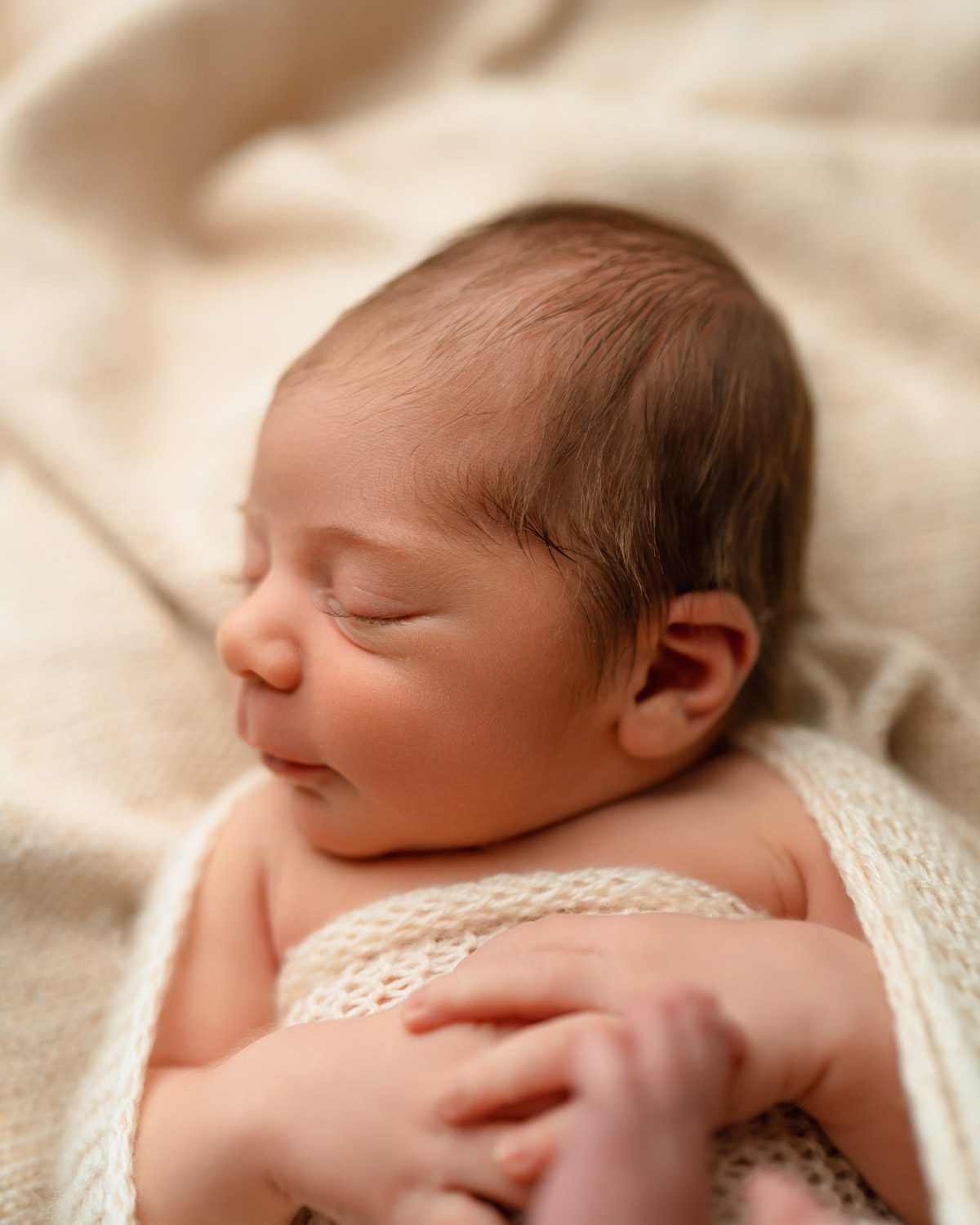 Newborn baby boy's facial features and eyelashes as he lays on his back swaddled in a cream knit wrap.