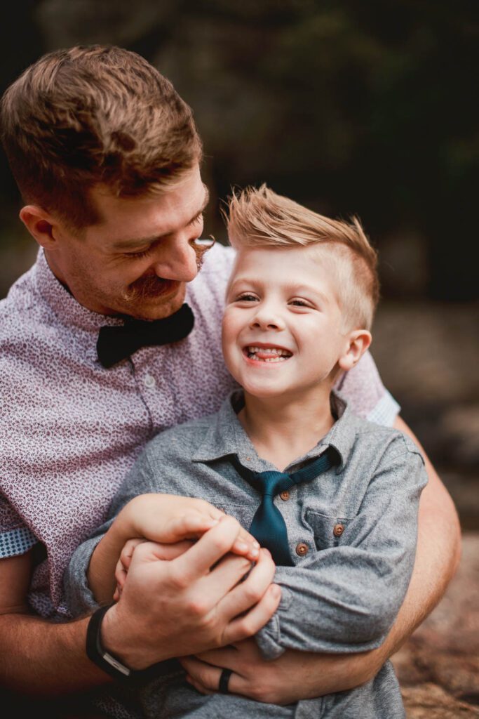 Dad holding his smiling older son close while standing beside broad rocks and flowing water.