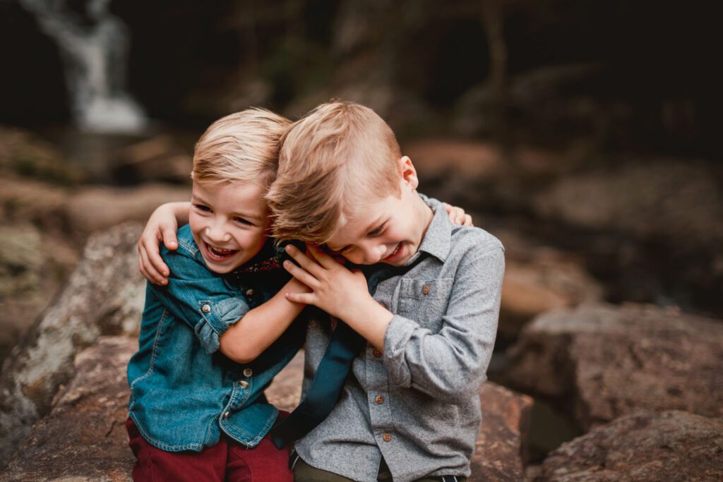 Two brothers laughing and hugging together on the shoals with a softly blurred waterfall behind them.