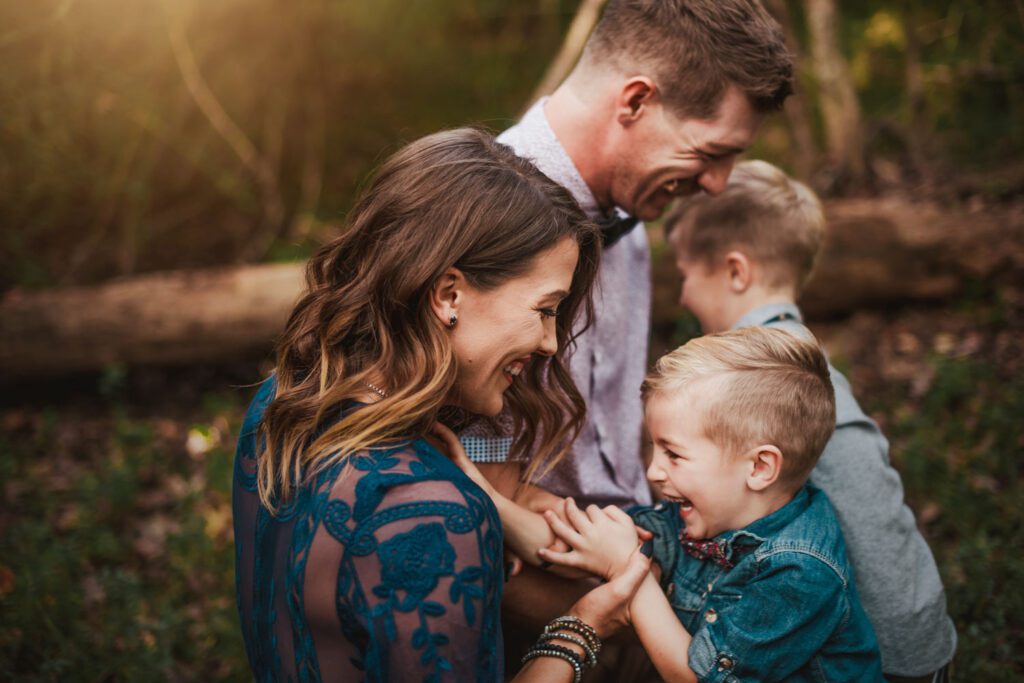 Mom and dad laughing with their two sons as the family cuddles closely together in warm woodland light.