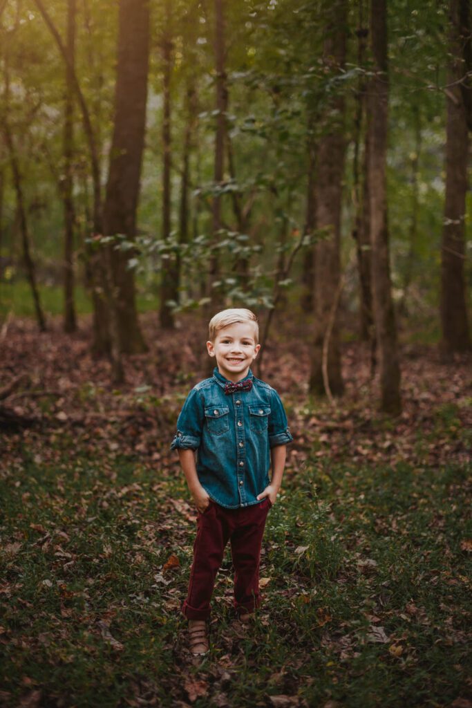 Young son in a denim shirt standing alone on a wooded trail surrounded by tall trees and fallen leaves.
