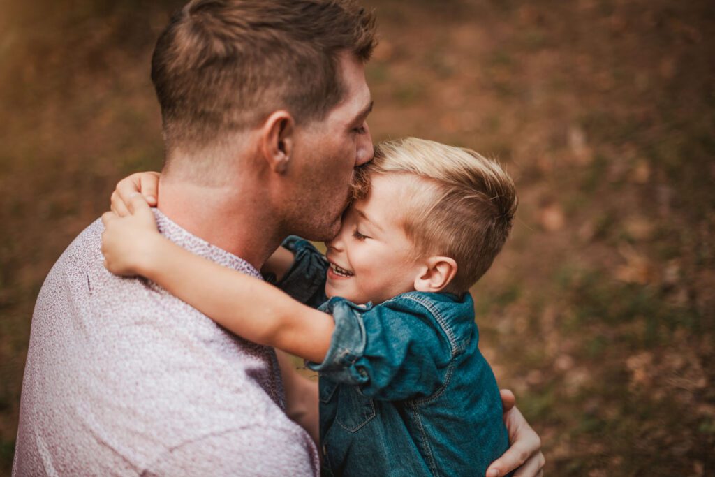Dad kissing his young son while holding him close against a softly blurred woodland background.