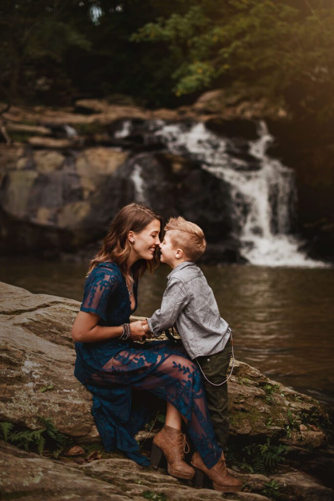 Mom kneeling on a rock while smiling nose-to-nose with her young son beside a cascading waterfall.
