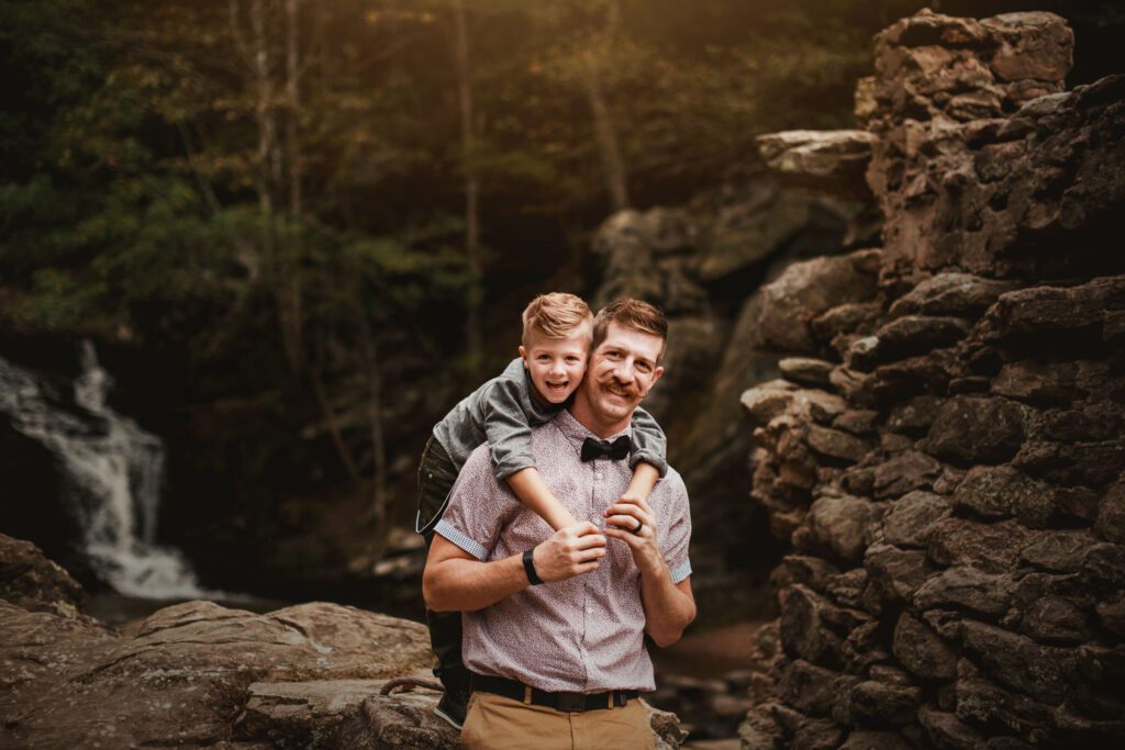 Dad carrying his older son on his shoulders beside large rocks and a wooded creek.