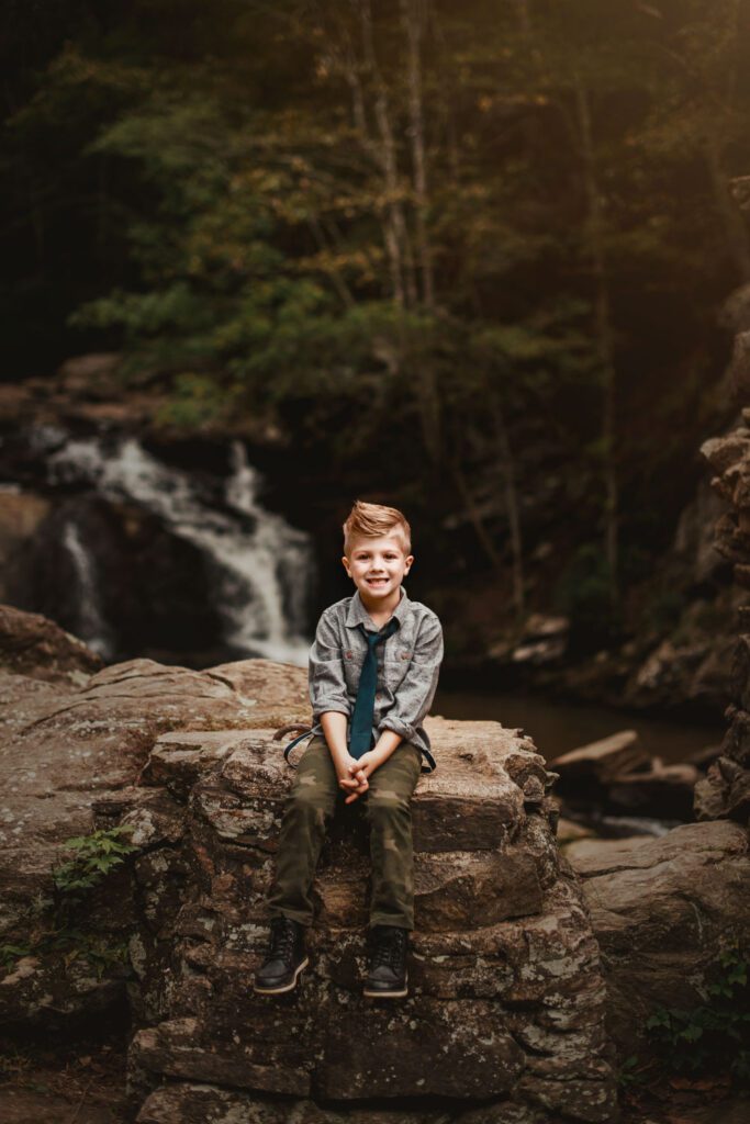 Young son in a gray shirt sitting on a large rock with wooded cliffs and flowing water behind him.
