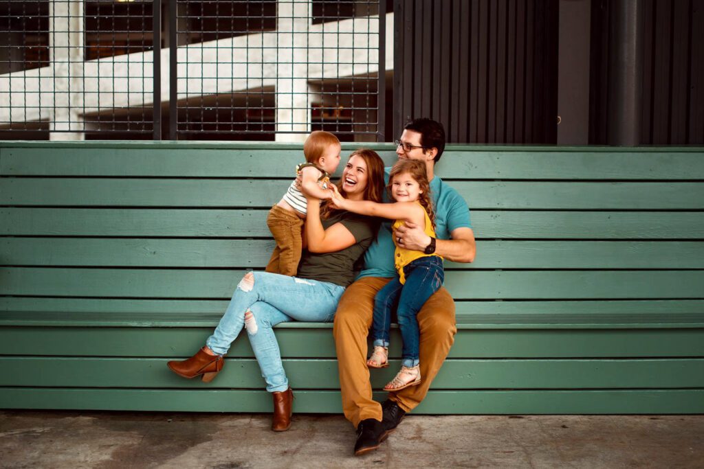 Mom, dad, daughter, and son sitting together on a teal bench with industrial steel windows behind them.