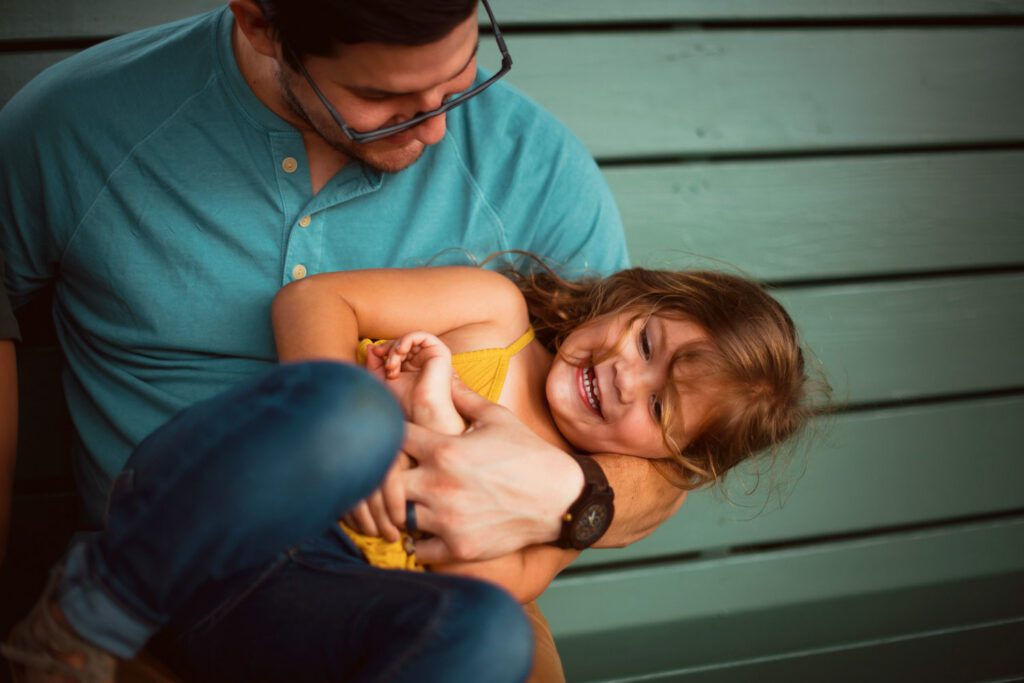 Dad holding his laughing daughter across his lap on a teal bench beneath gridded steel-framed windows.