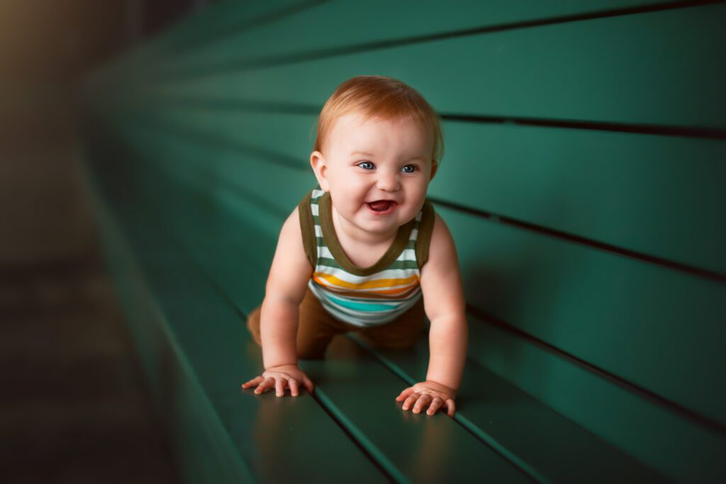 Smiling baby boy in a striped shirt crawling along a teal bench 