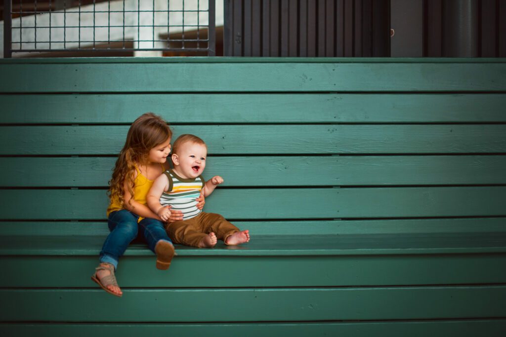 Siblings sitting together on a teal bench with large gridded steel windows in background