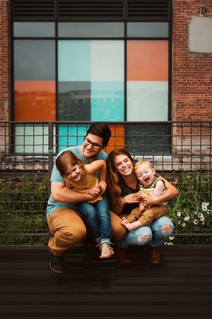Parents and siblings cuddling outside with Ponce City Market colorful stained-glass-style windows in background in Atlanta, GA