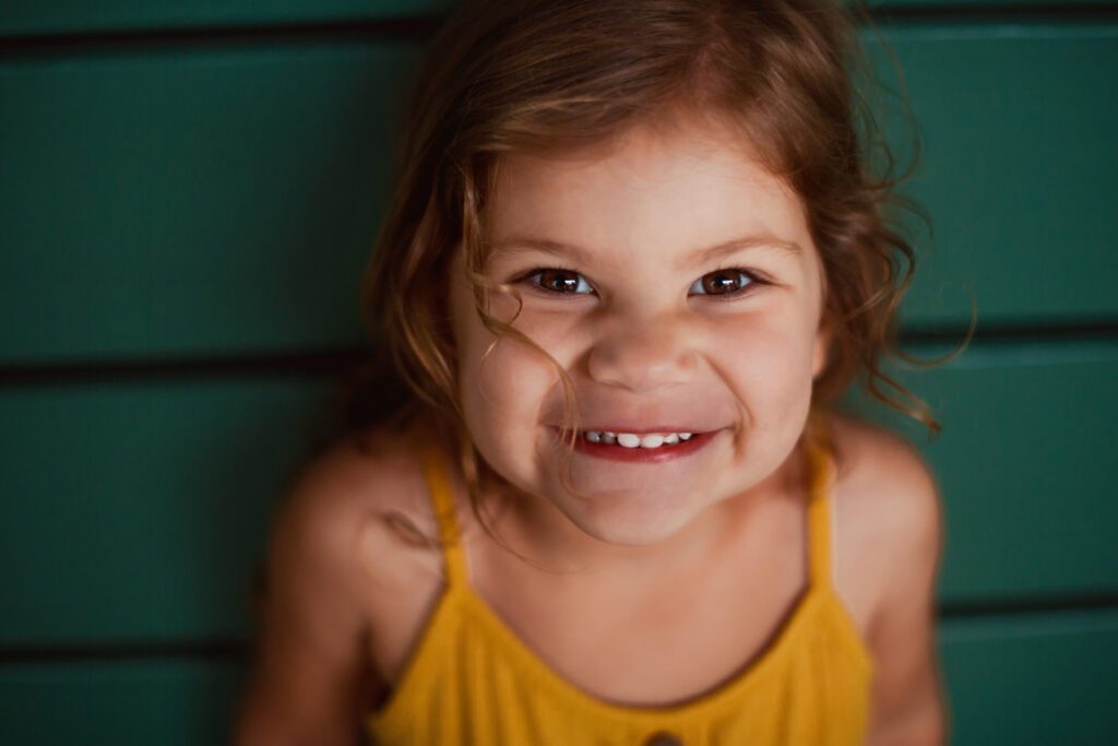 Close-up portrait of a smiling young girl in a mustard top against a softly blurred teal background.