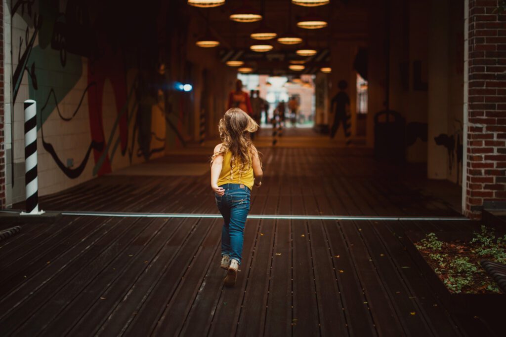 Young girl in a mustard top walking through the warm-lit entrance breezeway beneath vintage orange pendant lights.