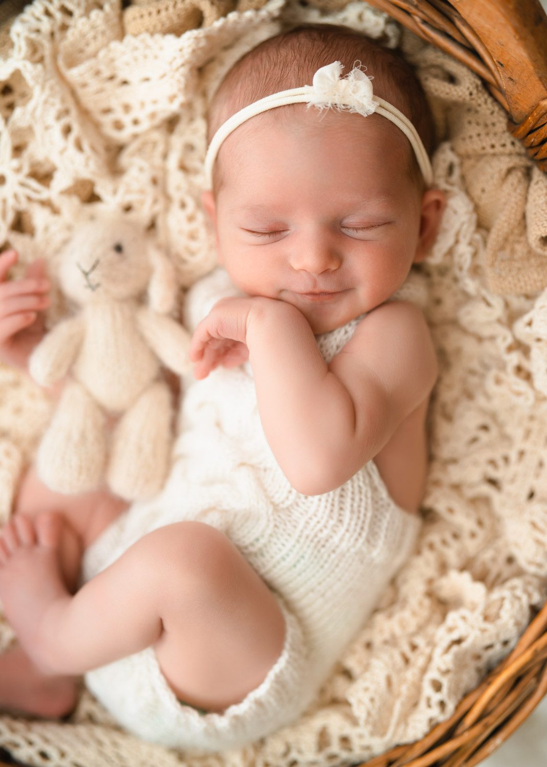 Smiling newborn baby girl holds a cream bunny stuffy wearing a white knit romper and laying in a wooden bowl.