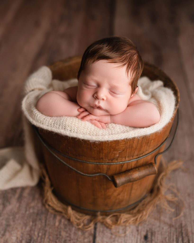 Wooden bucket pail with a newborn baby boy with red hair placed inside on a white blanket rests his head on his hands. 