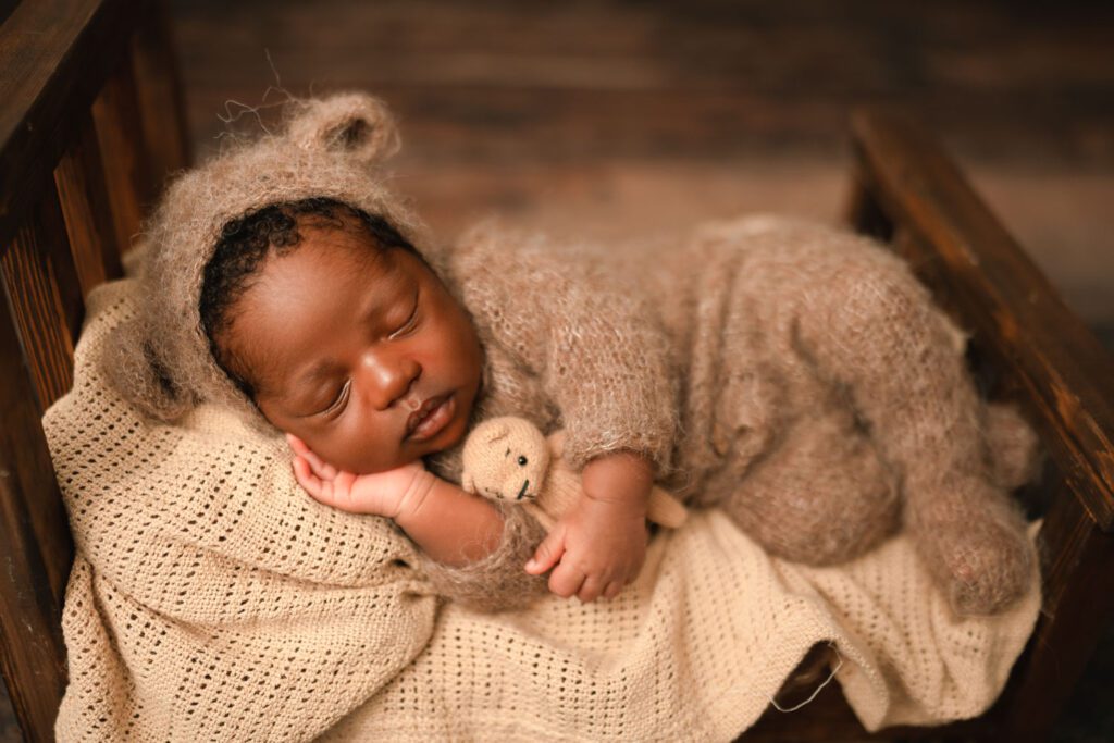 Newborn boy wears a brown knit outfit and matching bonnet with bear ears while holding a bear stuffy laying on a wooden bed. 