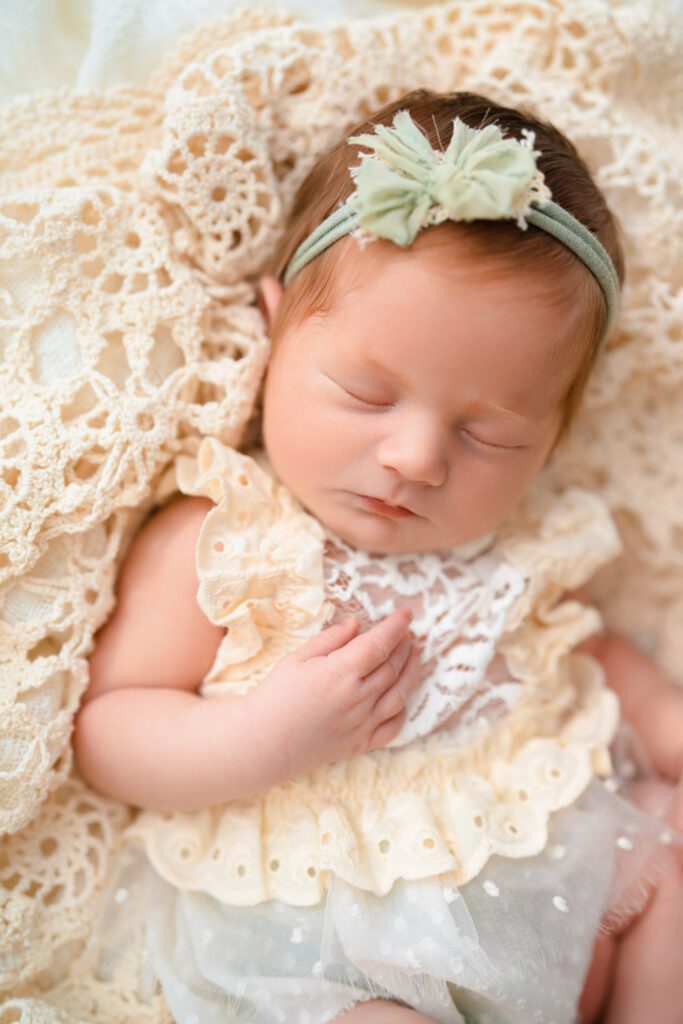Redhead newborn wears a lace newborn dress with a green bow headband in her hair as she lays on a vintage crochet blanket. 
