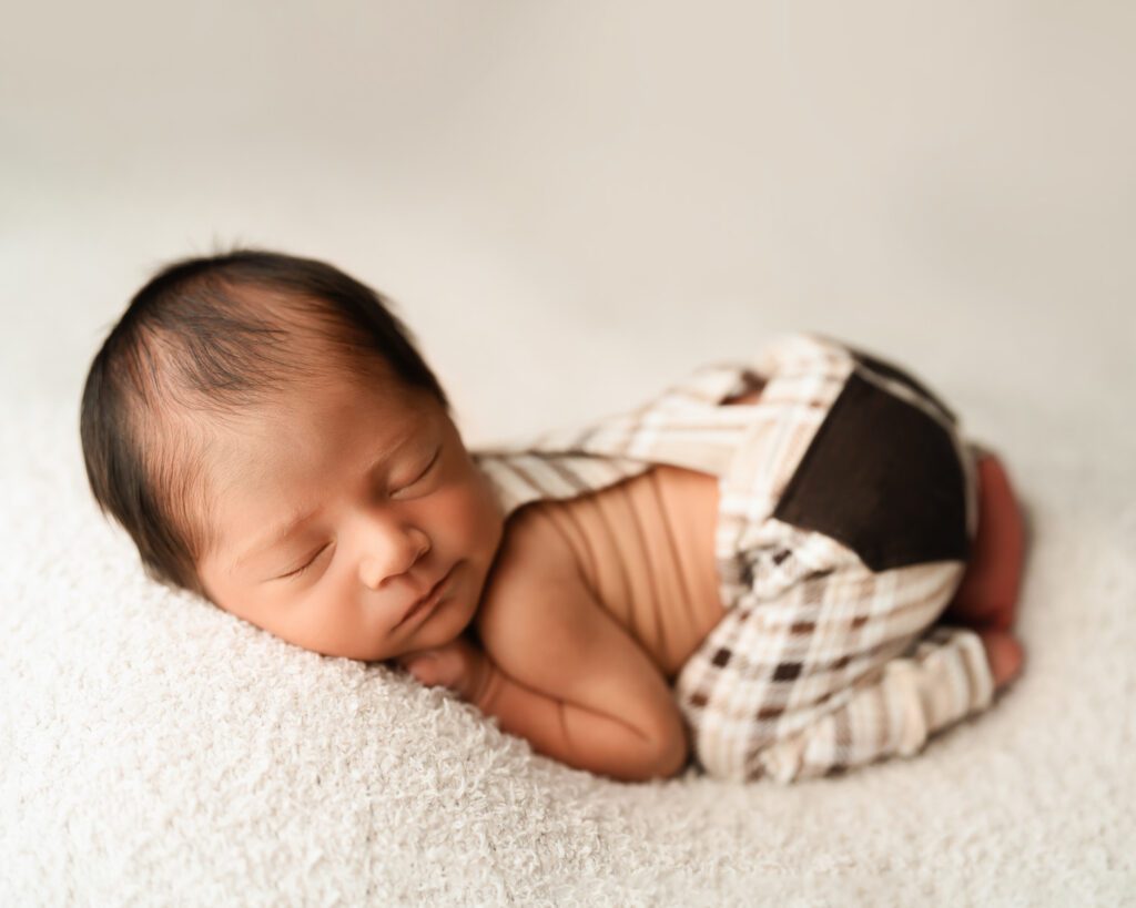 Newborn baby boy wearing white and brown plaid checkered suspender pants laying on a plus cream blanket backdrop. 