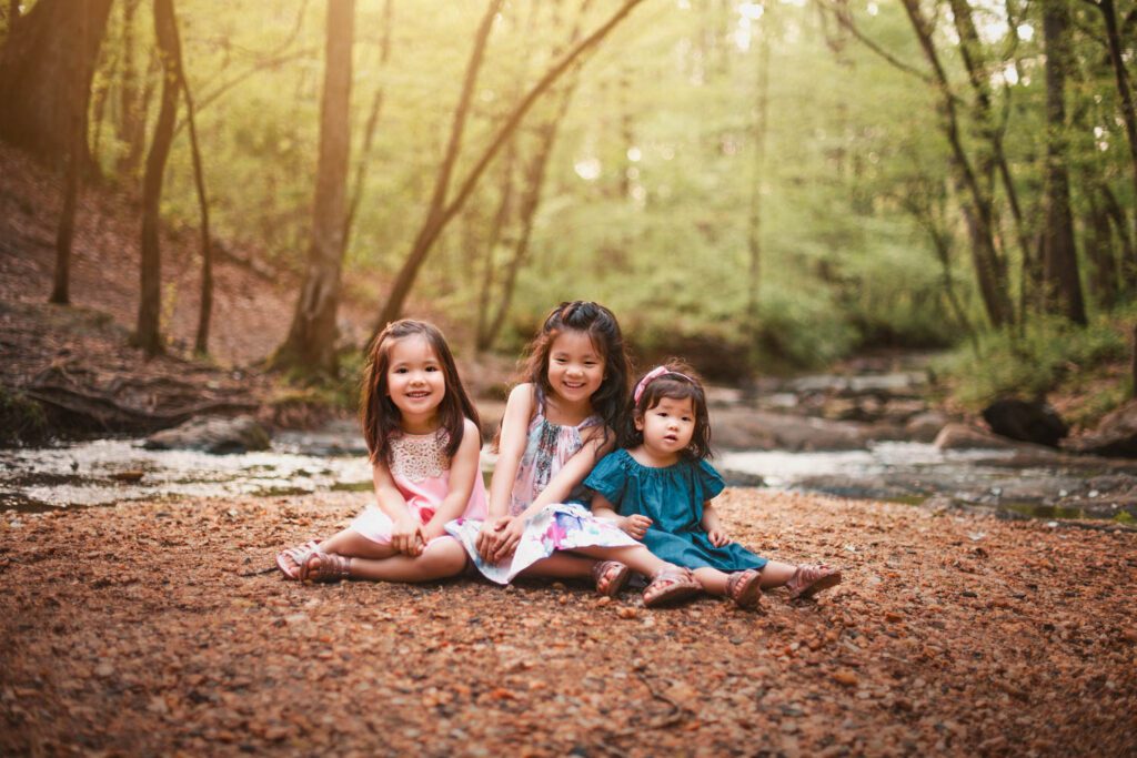 Three sisters sitting together beside Vickery Creek with the wooded creek bed in the background in Roswell, GA.