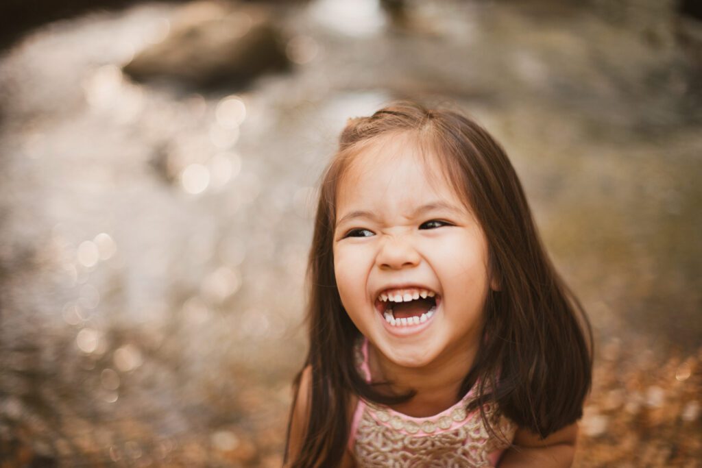 Close-up portrait of a laughing daughter with softly blurred creek water sparkling behind her.