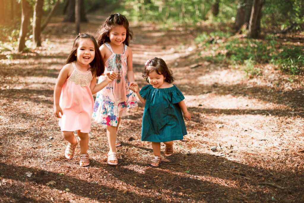 Three sisters walking together along a wooded trail surrounded by fallen leaves and warm sunlight.