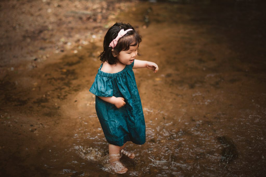 Young daughter in a teal dress wading through shallow creek water with her arms slightly raised.