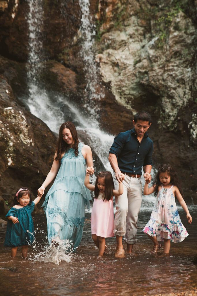 Parents walking hand in hand with their three daughters through shallow water beneath a rocky waterfall.