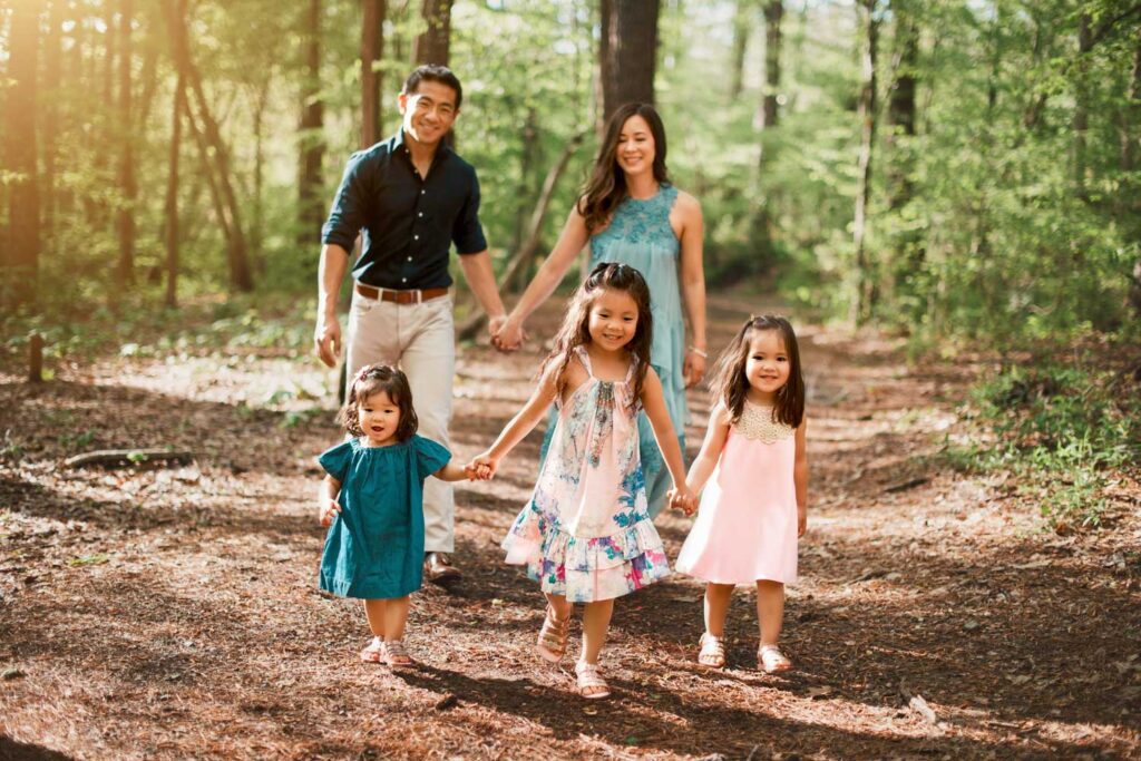 Parents walking with their three daughters along a wooded trail with warm sunlight filtering through the trees.