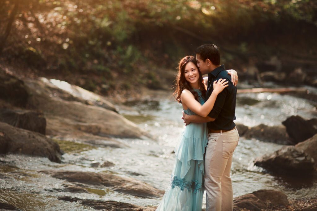 Mom and dad embracing beside a rocky creek with softly flowing water and wooded banks behind them.